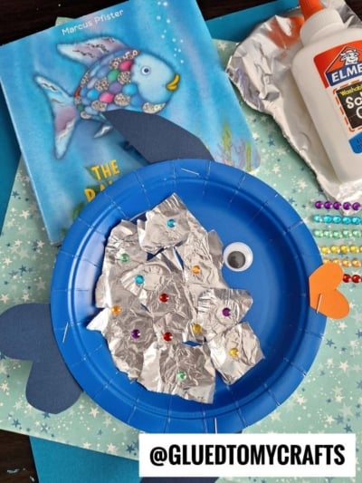A blue paper plate aluminum foil fish is decorated with sparkling foil scales, sequins, a large googly eye, and colorful paper fins. The book The Rainbow Fish and craft supplies like glue and beads are placed close by.