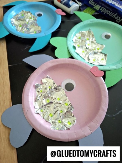 Three colorful paper plate fish crafts are shown, each with foil scales, a large googly eye, and cut-out paper fins and tails. The fish are on a table with craft supplies nearby. Text reads @GLUEDTOMYCRAFTS.