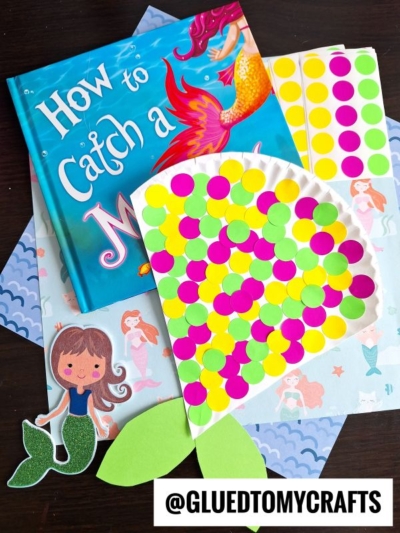 A colorful mermaid-themed craft setup features the book How to Catch a Mermaid, dot stickers on a shell outline, mermaid-themed paper, a paper mermaid cutout, and a coding label reading @GLUETOMYCRAFTS. Perfect for creative crafts!.