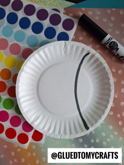 A white paper plate with a black curved line, colorful dot stickers, a black marker, and a handle with @GLUEDTOMYCRAFTS written on it, all arranged on a textured surface.