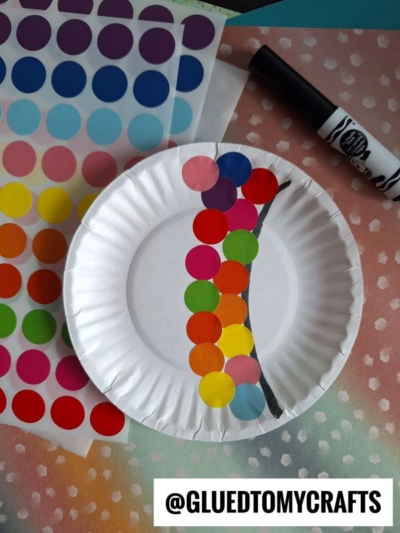 A white paper plate with a line drawn on it, decorated with colorful round stickers forming a caterpillar. Sheets of stickers and a black marker are nearby. @GLUEDTOMYCRAFTS is written at the bottom.
