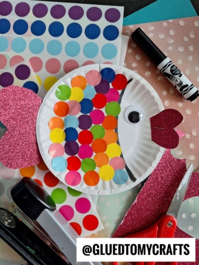 A paper plate fish craft decorated with colorful dot stickers, a large googly eye, and pink glitter heart shapes for fins, surrounded by craft supplies like markers, scissors, a stapler, and sticker sheets.