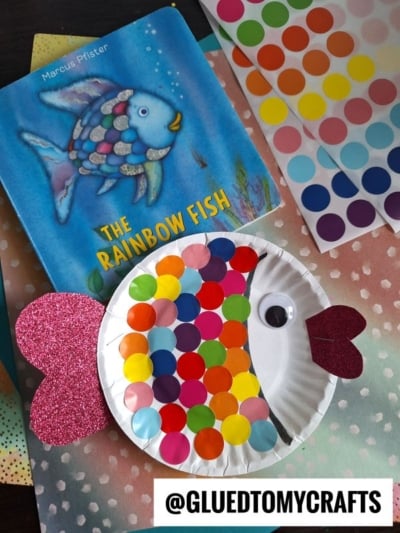 A children’s book, The Rainbow Fish, sits beside a paper plate fish craft covered in colorful dot stickers with a googly eye and glittery pink fins. An @GLUEDTOMYCRAFTS handle is visible at the bottom.