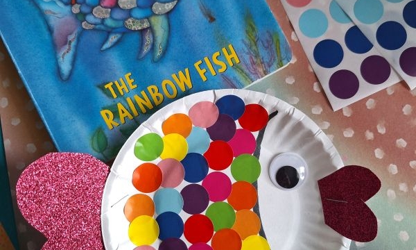A children’s book, The Rainbow Fish, sits beside a paper plate fish craft covered in colorful dot stickers with a googly eye and glittery pink fins. An @GLUEDTOMYCRAFTS handle is visible at the bottom.