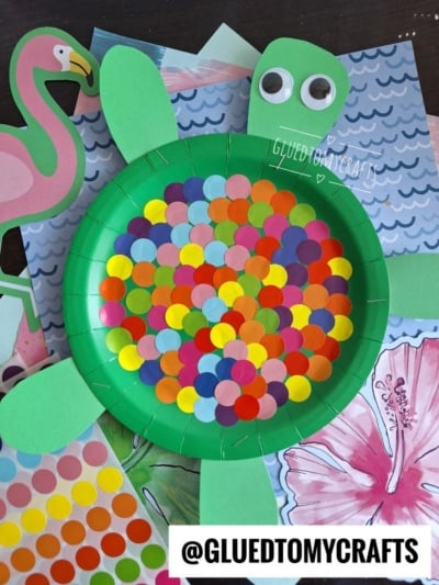 A green paper plate turtle craft with colorful dot stickers on its shell, green paper limbs, and googly eyes, placed on top of bright, tropical-themed patterned paper. @GLUEDTOMYCRAFTS is written at the bottom.