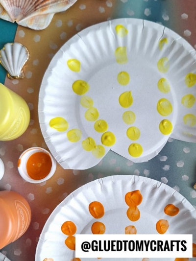 Two paper plates with yellow and orange paint dots are on a colorful surface with seashells and bottles of paint nearby. The handle @GLUETOMYCRAFTS appears in bold text at the bottom.