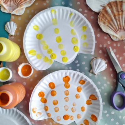 Two paper plates painted with yellow and orange dots to resemble seashells, surrounded by paint bottles, a pair of scissors, real seashells, and colorful paper. @GLUEDTOMYCRAFTS is written at the bottom.