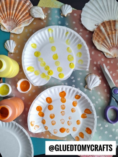 Two paper plates painted with yellow and orange dots to resemble seashells, surrounded by paint bottles, a pair of scissors, real seashells, and colorful paper. @GLUEDTOMYCRAFTS is written at the bottom.