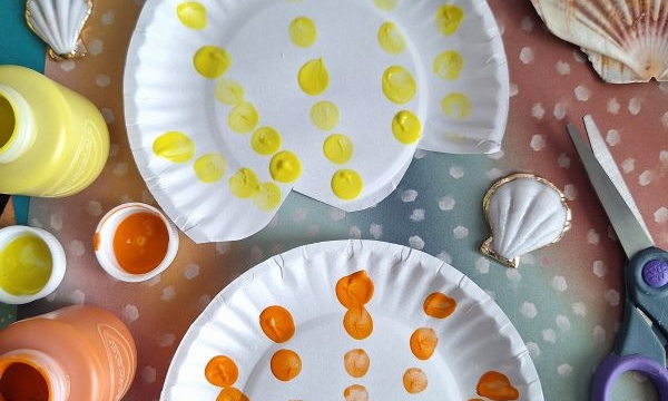 Two paper plates painted with yellow and orange dots to resemble seashells, surrounded by paint bottles, a pair of scissors, real seashells, and colorful paper. @GLUEDTOMYCRAFTS is written at the bottom.