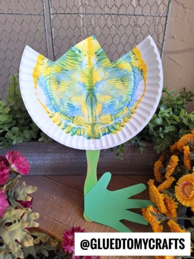 A vibrant PAINT SPLAT CRAFTS project: a paper plate painted yellow, blue, and green is folded to resemble a flower, with a green craft stick stem and a cut-out green paper hand as leaves, displayed among artificial flowers.