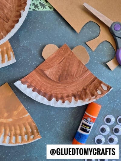 A paper plate painted brown with cut-out ears, a glue stick, googly eyes, scissors, and craft paper on a table. The project appears to be a bear craft in progress. @GLUEDTOMYCRAFTS text at the bottom.