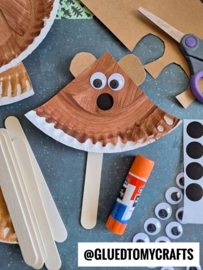 A bear craft made from a painted paper plate, googly eyes, and a popsicle stick, surrounded by craft supplies like glue, scissors, extra sticks, and googly eyes, on a blue surface.