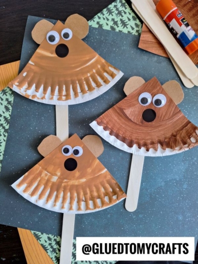 Three DIY bear crafts made from painted coffee filters, googly eyes, paper ears, and craft sticks are displayed on a table with craft supplies in the background. The text @GLUEDTOMYCRAFTS appears at the bottom.