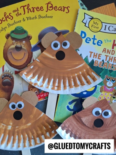 Three paper plate bear crafts with googly eyes and brown noses are displayed on top of children’s books, including “Goldilocks and the Three Bears” and “Pete the Cat and the Three Bears.”.