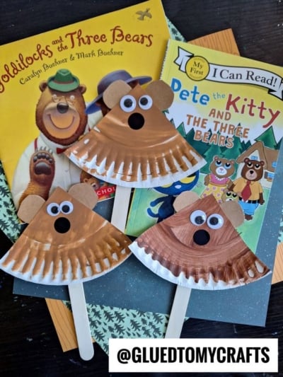 Three bear crafts made from paper plates, googly eyes, and craft sticks are displayed on top of the books Goldilocks and the Three Bears and Pete the Kitty and the Three Bears. A @GLUEDTOMYCRAFTS watermark is shown.