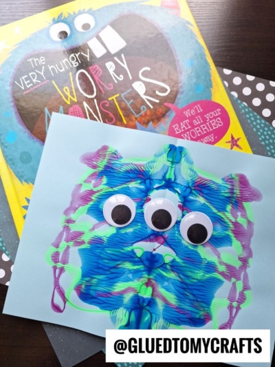 A children’s book titled The Very Hungry Worry Monsters and a colorful paint splat craft paper monster with three large googly eyes. The craft is made from paint blots on blue paper. Text in the corner reads @GLUEDTOMYCRAFTS.