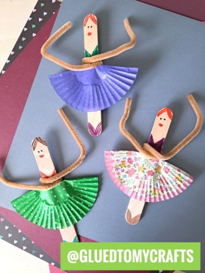 Three ballerina popsicle stick craft ideas feature wooden sticks, cupcake liner skirts, and pipe cleaner arms, all arranged on colorful paper. Each has a unique drawn face and hairstyle. The handle @GLUETOMYCRAFTS appears in the corner.