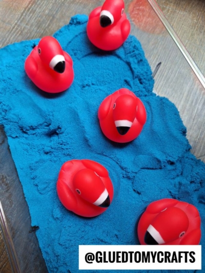 Five red rubber flamingo toys sit on textured blue kinetic sand inside a clear container. A white label at the bottom reads @GLUEDTOMYCRAFTS.
