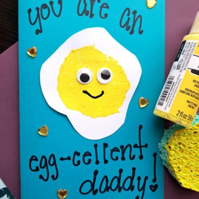 A handmade Father’s Day card features a smiling fried egg with googly eyes on a blue background, decorated using sponge painting ideas for kids. The card reads, “You are an egg-cellent daddy!” Hearts and craft supplies surround it.