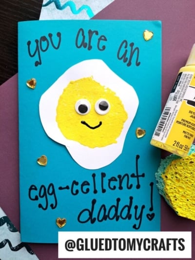 A handmade Father’s Day card features a smiling fried egg with googly eyes on a blue background, decorated using sponge painting ideas for kids. The card reads, “You are an egg-cellent daddy!” Hearts and craft supplies surround it.