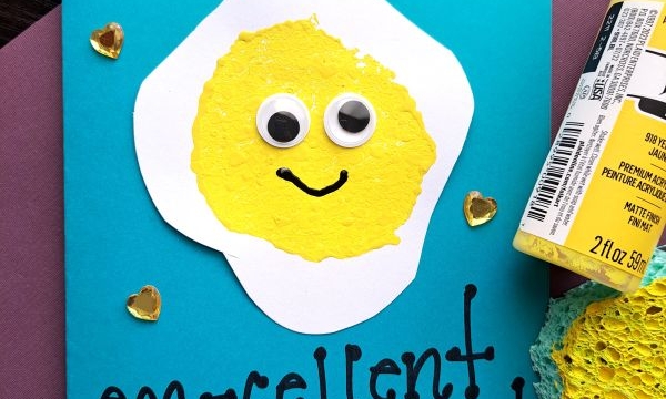 A handmade Father’s Day card features a smiling fried egg with googly eyes on a blue background, decorated using sponge painting ideas for kids. The card reads, “You are an egg-cellent daddy!” Hearts and craft supplies surround it.