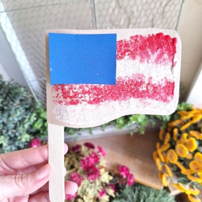 A hand holds a craft resembling an American flag, made with a popsicle stick handle, red and white textured stripes, and a blue square in the top left corner. Flowers and greenery are visible in the background.