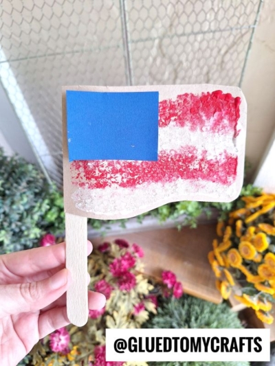 A hand holds a craft resembling an American flag, made with a popsicle stick handle, red and white textured stripes, and a blue square in the top left corner. Flowers and greenery are visible in the background.