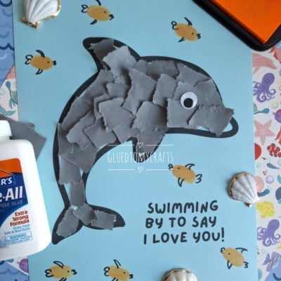 A childrens craft project features a dolphin made of torn gray paper pieces on blue paper, surrounded by drawn fish, seashells, glue, and the text Swimming by to say I love you! with the @GLUEDTOMYCRAFTS tag.