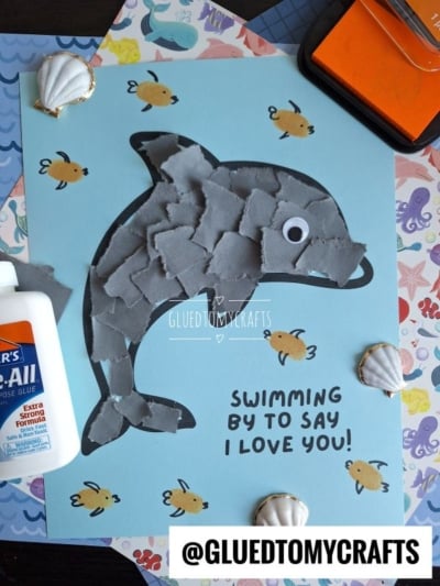 A childrens craft project features a dolphin made of torn gray paper pieces on blue paper, surrounded by drawn fish, seashells, glue, and the text Swimming by to say I love you! with the @GLUEDTOMYCRAFTS tag.
