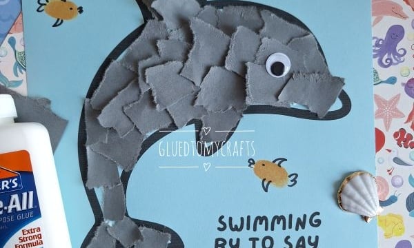 A childrens craft project features a dolphin made of torn gray paper pieces on blue paper, surrounded by drawn fish, seashells, glue, and the text Swimming by to say I love you! with the @GLUEDTOMYCRAFTS tag.