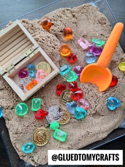 A wooden chest filled with gold coins and colorful plastic gems sits on kinetic sand. More coins and gems are scattered around, along with an orange plastic scoop. The text @GLUEDTOMYCRAFTS appears at the bottom.