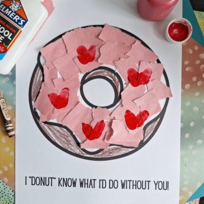 A donut coloring page with pink paper pieces and red hearts glued on top, surrounded by a bottle of glue, red paint, a paintbrush, a crayon, and colorful papers. Text on the page reads “I ‘donut’ know what I’d do without you!”.