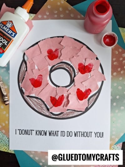 A donut coloring page with pink paper pieces and red hearts glued on top, surrounded by a bottle of glue, red paint, a paintbrush, a crayon, and colorful papers. Text on the page reads “I ‘donut’ know what I’d do without you!”.