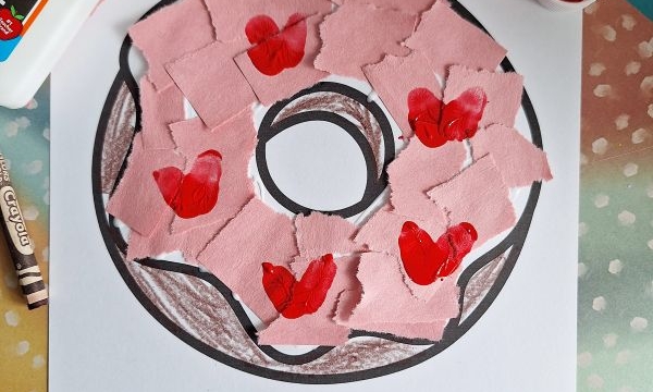 A donut coloring page with pink paper pieces and red hearts glued on top, surrounded by a bottle of glue, red paint, a paintbrush, a crayon, and colorful papers. Text on the page reads “I ‘donut’ know what I’d do without you!”.