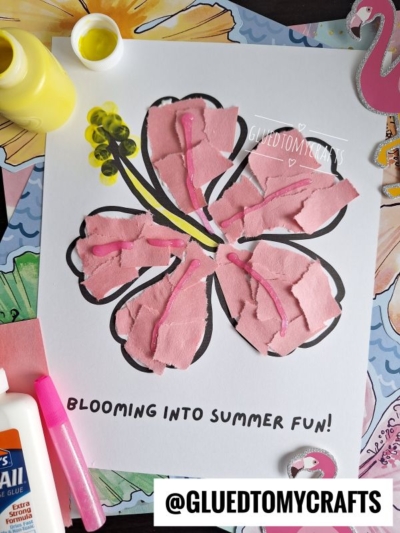 A craft project of a hibiscus flower made with torn pink paper pieces glued onto a printed outline, with yellow and black details. Supplies like glue, yellow paint, and a brush are nearby. Text reads “BLOOMING INTO SUMMER FUN!.