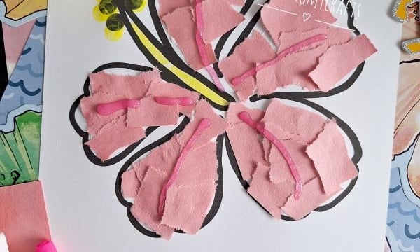 A craft project of a hibiscus flower made with torn pink paper pieces glued onto a printed outline, with yellow and black details. Supplies like glue, yellow paint, and a brush are nearby. Text reads “BLOOMING INTO SUMMER FUN!.