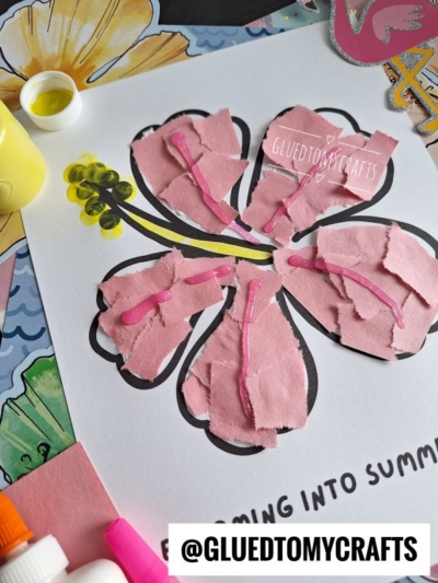 A flower craft made with torn pink paper pieces glued onto a black outline of a flower, with pink glitter glue accents. Art supplies and colorful paper are scattered around. Text at the bottom reads @GLUETOMYCRAFTS.