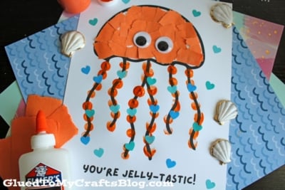 A handmade jellyfish craft on white paper, with an orange torn-paper body, googly eyes, wavy orange ribbon tentacles, and blue heart decorations. The text below reads, YOURE JELLY-TASTIC! Various seashells and craft materials surround it.