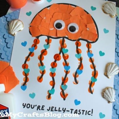 A handmade jellyfish craft on white paper, with an orange torn-paper body, googly eyes, wavy orange ribbon tentacles, and blue heart decorations. The text below reads, YOURE JELLY-TASTIC! Various seashells and craft materials surround it.