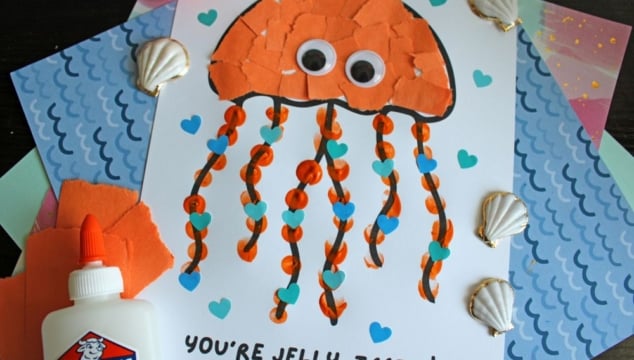 A handmade jellyfish craft on white paper, with an orange torn-paper body, googly eyes, wavy orange ribbon tentacles, and blue heart decorations. The text below reads, YOURE JELLY-TASTIC! Various seashells and craft materials surround it.