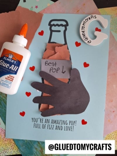 A handmade Father’s Day card featuring a torn paper soda bottle with a hand cutout holding it, hearts, and a note reading “Best Pop!” Supplies include glue and craft materials. Text at the bottom reads “You’re an amazing pop! Full of fizz and love!”.