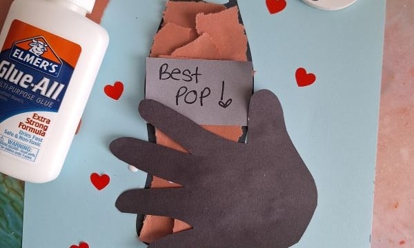 A handmade Father’s Day card featuring a torn paper soda bottle with a hand cutout holding it, hearts, and a note reading “Best Pop!” Supplies include glue and craft materials. Text at the bottom reads “You’re an amazing pop! Full of fizz and love!”.