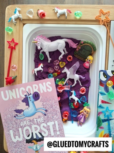 A sensory tray with purple playdough, unicorn figurines, a fairy door, colorful gems, star wands, a small doll, and a pink book titled Unicorns Are The Worst! lies next to a @gluedtomycrafts sign.