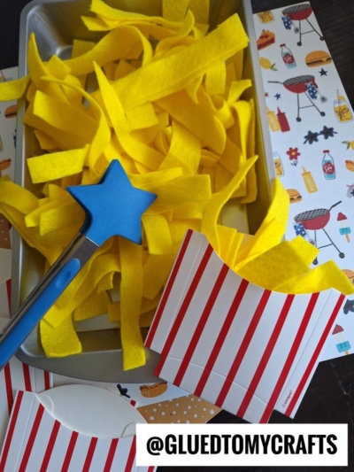 A tray filled with yellow felt strips resembling French fries, a blue star-tipped spatula, and two red-striped paper fry containers—perfect for story time sensory bin ideas. Background features barbecue-themed illustrations. @GLUEDTOMYCRAFTS is at the bottom.