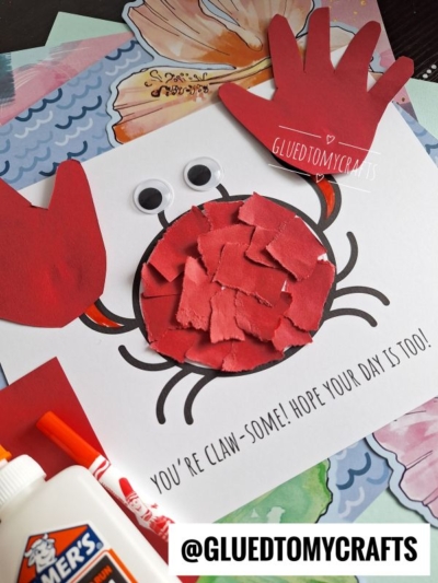 A childrens craft of a red crab made from torn paper pieces, googly eyes, and red paper claws on a white sheet with the text Youre Claw-some! Hope your day is too! Craft supplies are nearby.