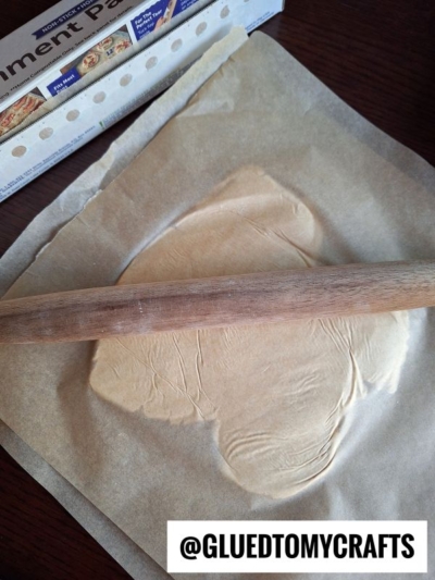 A rolling pin flattens dough on parchment paper, with a box of parchment paper visible in the background. Text at the bottom reads @GLUETOMYCRAFTS.