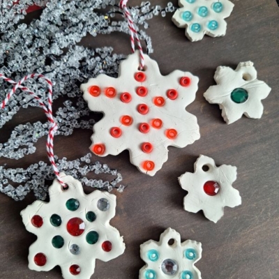 Handmade white clay snowflake ornaments with colorful rhinestones and string, displayed on a dark surface with frosted decorative branches. A label reads @GLUEDTOMYCRAFTS at the bottom.