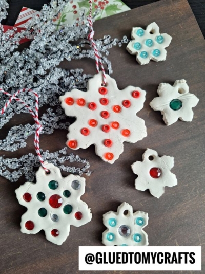Handmade white clay snowflake ornaments with colorful rhinestones and string, displayed on a dark surface with frosted decorative branches. A label reads @GLUEDTOMYCRAFTS at the bottom.