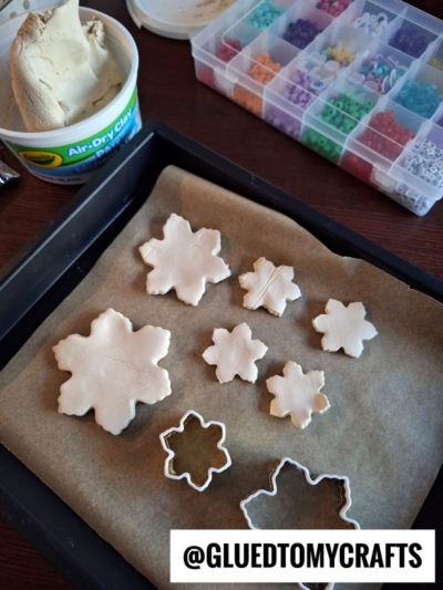 Air-dry clay snowflakes are cut out on parchment paper in a baking tray, with a snowflake-shaped cutter, a tub of clay, and a box of colorful beads visible nearby. The text @GLUEDTOMYCRAFTS is at the bottom.