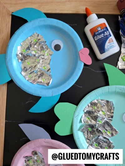 Three paper plate fish crafts with blue, green, and pink plates, decorated with tin foil, googly eyes, and colored paper fins, next to a bottle of glue. Text reads @GLUEDTOMYCRAFTS.